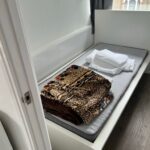 Property for sale by Auction A small bedroom with a single bed, featuring a gray mattress. There's a stack of folded white sheets and a patterned blanket on the bed. A window with a partially open curtain lets in natural light, and the room has wooden flooring. Presented by Palace Auctions