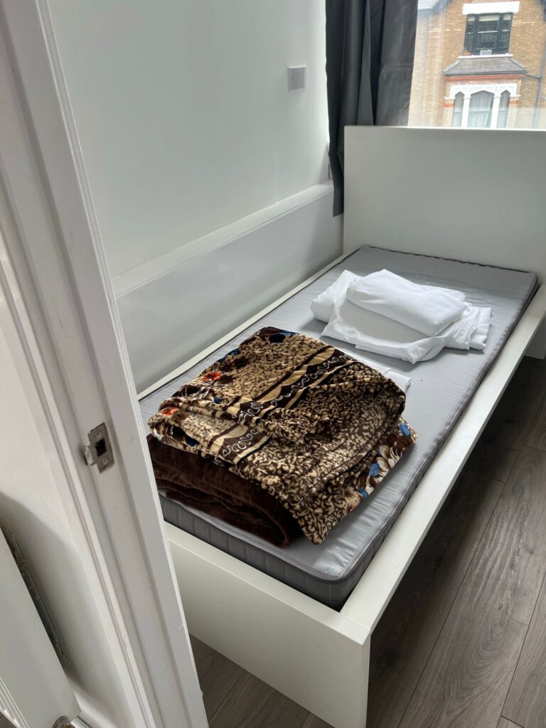 Property for sale by Auction A small bedroom with a single bed, featuring a gray mattress. There's a stack of folded white sheets and a patterned blanket on the bed. A window with a partially open curtain lets in natural light, and the room has wooden flooring. Presented by Palace Auctions