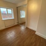Property for sale by Auction An empty room with light brown wooden flooring and cream-colored walls. There are two windows with white frames, a radiator beneath one window, and a mirrored sliding closet door on one side. Presented by Palace Auctions