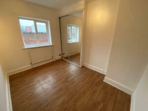 Property for sale by Auction An empty room with light brown wooden flooring and cream-colored walls. There are two windows with white frames, a radiator beneath one window, and a mirrored sliding closet door on one side. Presented by Palace Auctions