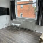 Property for sale by Auction A minimalist room with two white folding chairs in front of a large window with gray curtains. The floor is wooden, and a flat-screen TV is mounted on the white wall. A brick building is visible through the window. Presented by Palace Auctions
