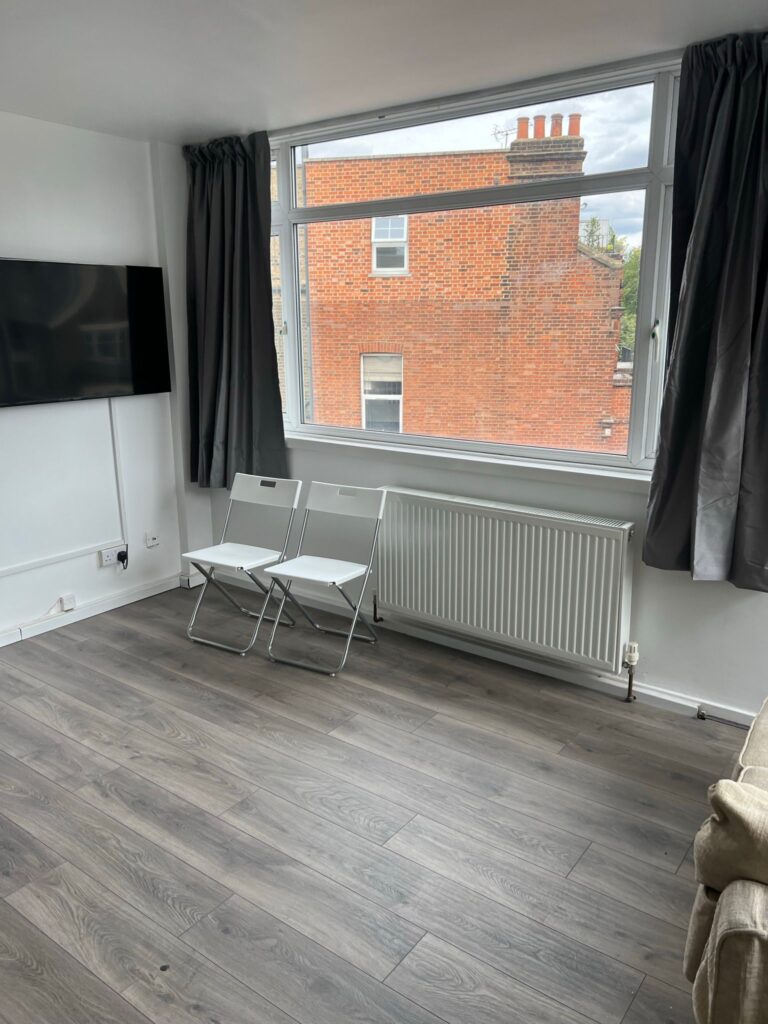 Property for sale by Auction A minimalist room with two white folding chairs in front of a large window with gray curtains. The floor is wooden, and a flat-screen TV is mounted on the white wall. A brick building is visible through the window. Presented by Palace Auctions