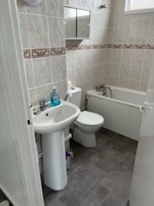 Property for sale by Auction A small bathroom with white and beige tiled walls. It features a pedestal sink with a liquid soap dispenser, a toilet, a bathtub, and a mirrored cabinet above the sink. The floor is covered with gray tiles. Presented by Palace Auctions