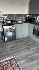 Property for sale by Auction A modern kitchen with gray cabinets, black tile backsplash, and wooden flooring. A washing machine and oven are integrated into the cabinetry. A colorful rug is partially visible on the floor. Various kitchen items are placed on the countertop. Presented by Palace Auctions