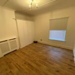 Property for sale by Auction An empty room with wooden floors, white walls, and a ceiling light fixture. There are built-in white wardrobes, a radiator cover, a window with a closed blind, and shelving on one side. Presented by Palace Auctions