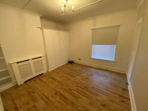 Property for sale by Auction An empty room with wooden floors, white walls, and a ceiling light fixture. There are built-in white wardrobes, a radiator cover, a window with a closed blind, and shelving on one side. Presented by Palace Auctions