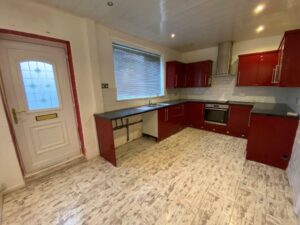 Property for sale by Auction A kitchen with red cabinets, a tiled floor, and white walls. It features an oven, stove, and a metallic sink under a large window. A door with a decorative glass panel is on the left. The room has modern lighting and a white painted ceiling. Presented by Palace Auctions