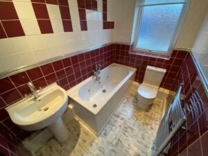 Property for sale by Auction A bathroom with a white sink, a bathtub, and a toilet. The walls are tiled with red and white tiles, and there's a frosted window. The floor has a light-colored wood pattern, and a metallic towel rack is mounted on the wall. Presented by Palace Auctions