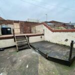 Property for sale by Auction An outdoor area with concrete ground and multi-level wooden decking. Stairs connect the levels, and a brick wall with barbed wire surrounds the space. A trash bin is visible near the top. The sky is overcast, creating a gloomy atmosphere. Presented by Palace Auctions
