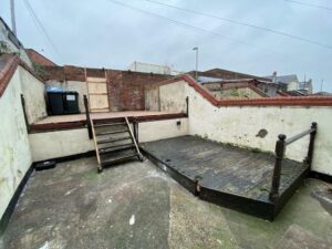 Property for sale by Auction An outdoor area with concrete ground and multi-level wooden decking. Stairs connect the levels, and a brick wall with barbed wire surrounds the space. A trash bin is visible near the top. The sky is overcast, creating a gloomy atmosphere. Presented by Palace Auctions
