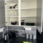 Property for sale by Auction Open kitchen cabinet showing stacked bowls, plates, glasses, and cooking pots. Below, on the countertop, are a kettle, toaster, and labeled canisters for sugar, coffee, and tea. The backsplash features gray tiles. Presented by Palace Auctions