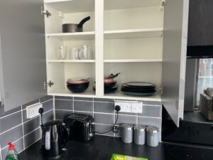 Property for sale by Auction Open kitchen cabinet showing stacked bowls, plates, glasses, and cooking pots. Below, on the countertop, are a kettle, toaster, and labeled canisters for sugar, coffee, and tea. The backsplash features gray tiles. Presented by Palace Auctions
