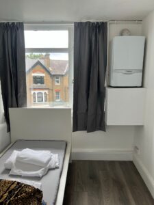 Property for sale by Auction A small room with a single bed and folded linens. A window with dark curtains shows a view of a brick house outside. A white boiler is mounted on the wall next to the window. The floor is dark wood. Presented by Palace Auctions