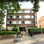 Property for sale by Auction A three-story apartment building with a brown and white exterior, surrounded by trees and shrubs. A blue bicycle is parked at the front near a bus stop, situated on a tree-lined street. Presented by Palace Auctions