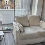 Property for sale by Auction A cozy room featuring a beige sofa with cushions, a sliding glass door leading to a small balcony, and a view of neighboring houses. The room has a wooden floor and a patterned rug. A white chair is partially visible on the left. Presented by Palace Auctions