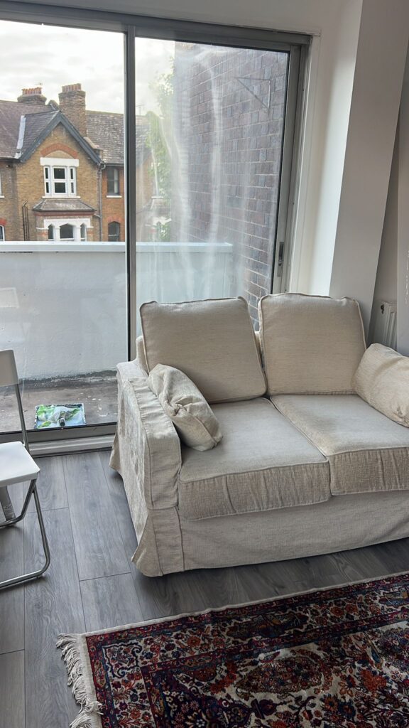 Property for sale by Auction A cozy room featuring a beige sofa with cushions, a sliding glass door leading to a small balcony, and a view of neighboring houses. The room has a wooden floor and a patterned rug. A white chair is partially visible on the left. Presented by Palace Auctions