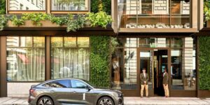 Property for sale by Auction A luxury car is parked in front of a modern building with a green, plant-covered facade, setting Mayfair's new benchmark. Two doormen stand at the entrance in uniform, as large windows reflect the thriving urban landscape of central London's luxury apartments. Presented by Palace Auctions