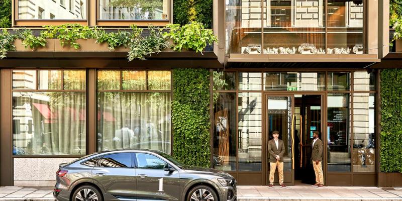 Property for sale by Auction A luxury car is parked in front of a modern building with a green, plant-covered facade, setting Mayfair's new benchmark. Two doormen stand at the entrance in uniform, as large windows reflect the thriving urban landscape of central London's luxury apartments. Presented by Palace Auctions