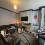 Property for sale by Auction A cluttered living room with a black leather sofa, a TV mounted on a blue and gray striped wall, and a gray carpet. Items are scattered on a small table and floor. A vacuum cleaner is visible in the foreground. Presented by Palace Auctions