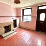 Property for sale by Auction A small, empty room with pink textured walls and a red geometric-patterned floor. It features a fireplace, a dark wooden door with a window, and a large window letting in natural light. An exposed light fixture hangs from the ceiling. Presented by Palace Auctions