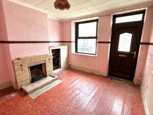 Property for sale by Auction A small, empty room with pink textured walls and a red geometric-patterned floor. It features a fireplace, a dark wooden door with a window, and a large window letting in natural light. An exposed light fixture hangs from the ceiling. Presented by Palace Auctions