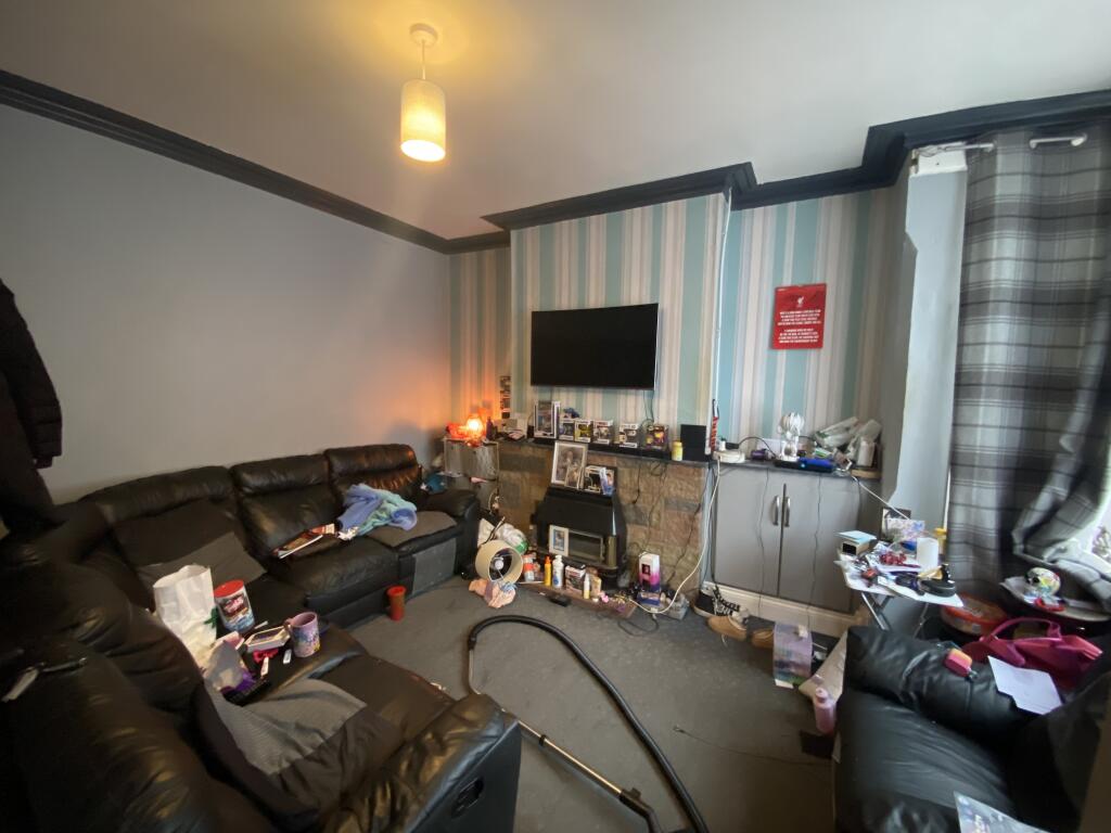 Property for sale by Auction A cluttered living room with a black leather sofa, a TV mounted on a blue and gray striped wall, and a gray carpet. Items are scattered on a small table and floor. A vacuum cleaner is visible in the foreground. Presented by Palace Auctions