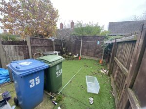 Property for sale by Auction A small fenced yard with artificial grass, fallen leaves, and scattered debris. Two large bins, one blue and one green, stand on the left. A clear plastic box and some tools are on the ground. There's a bare tree and some orange traffic cones in the background. Presented by Palace Auctions