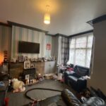 Property for sale by Auction A cluttered living room with striped wallpaper, a wall-mounted TV, and a large window with curtains. The room has two black leather sofas, with various items scattered around, including a vacuum and personal belongings. A ceiling light is on. Presented by Palace Auctions