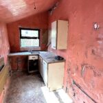 Property for sale by Auction A small, worn-out kitchen with peeling red walls, a dirty floor, and an empty cabinet. The room has a single window and an old sink. Overall, the space looks neglected and in need of renovation. Presented by Palace Auctions
