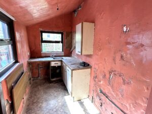 Property for sale by Auction A small, worn-out kitchen with peeling red walls, a dirty floor, and an empty cabinet. The room has a single window and an old sink. Overall, the space looks neglected and in need of renovation. Presented by Palace Auctions