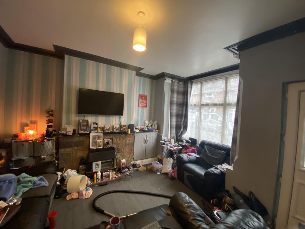 Property for sale by Auction A cluttered living room with striped wallpaper, a wall-mounted TV, and a large window with curtains. The room has two black leather sofas, with various items scattered around, including a vacuum and personal belongings. A ceiling light is on. Presented by Palace Auctions