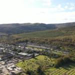 Property for sale by Auction Aerial view of a small town surrounded by lush green landscapes and hills under a partly cloudy sky. Residential buildings and patches of forested areas are visible, along with open fields and a cemetery in the foreground. Presented by Palace Auctions