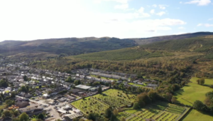 Property for sale by Auction Aerial view of a small town surrounded by lush green landscapes and hills under a partly cloudy sky. Residential buildings and patches of forested areas are visible, along with open fields and a cemetery in the foreground. Presented by Palace Auctions