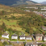 Property for sale by Auction Aerial view of a residential area with houses bordering a large green hillside. The landscape shows a blend of greenery and urban development, with mountains visible in the background under a clear sky. Presented by Palace Auctions