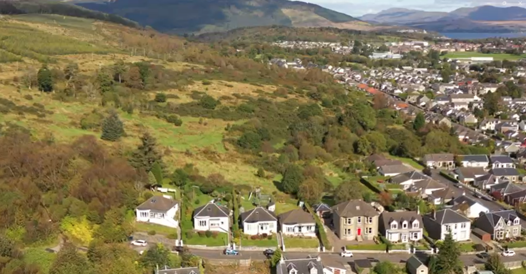 Property for sale by Auction Aerial view of a residential area with houses bordering a large green hillside. The landscape shows a blend of greenery and urban development, with mountains visible in the background under a clear sky. Presented by Palace Auctions