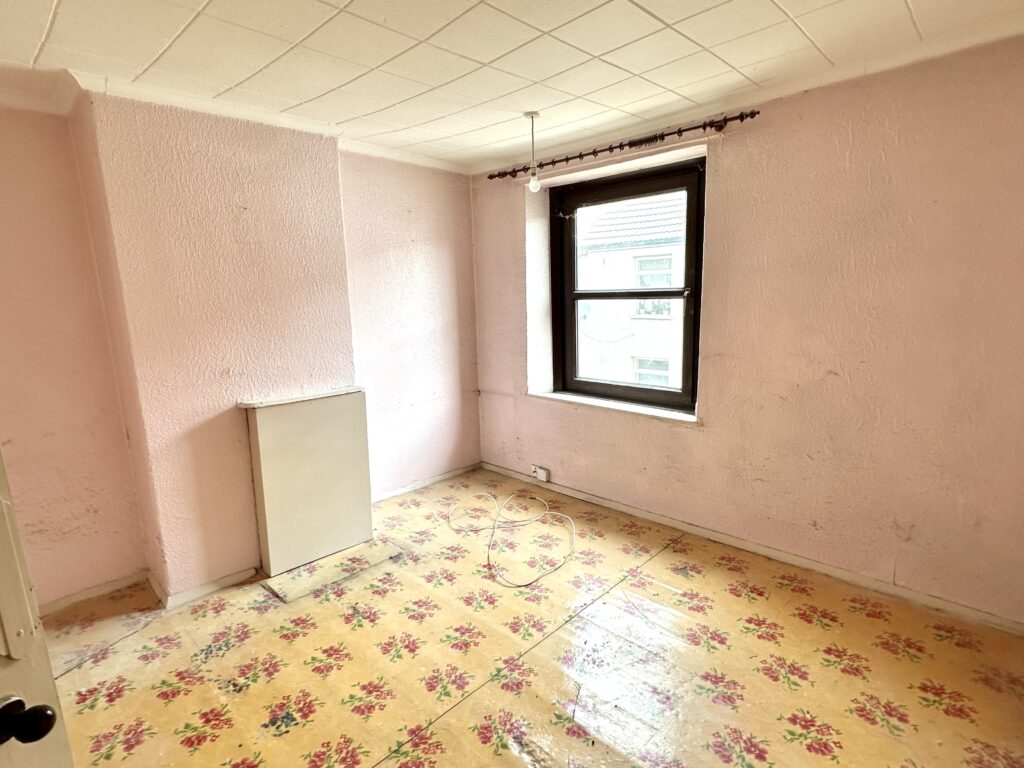 Property for sale by Auction An empty room with light pink walls, a single window with a black frame, and a small white radiator in the corner. The floor is covered with a yellow floral-patterned carpet, and a single bulb hangs from the ceiling. Presented by Palace Auctions