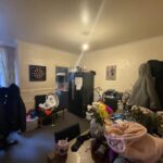 Property for sale by Auction A cluttered room with a table covered in clothes and other items. A black refrigerator stands in the corner, with a dartboard hanging on the wall. Two large coats hang on the back of chairs. A bouquet of colorful flowers is on the table. Presented by Palace Auctions
