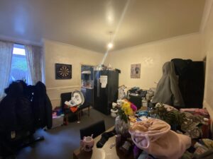 Property for sale by Auction A cluttered room with a table covered in clothes and other items. A black refrigerator stands in the corner, with a dartboard hanging on the wall. Two large coats hang on the back of chairs. A bouquet of colorful flowers is on the table. Presented by Palace Auctions