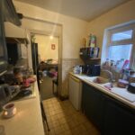 Property for sale by Auction A narrow kitchen with dark cabinets and tiled floor. Dishes are piled in a drying rack beside a sink. Small appliances like a microwave and kettle are on the counters. There's a doorway leading to a cluttered adjacent room. Presented by Palace Auctions
