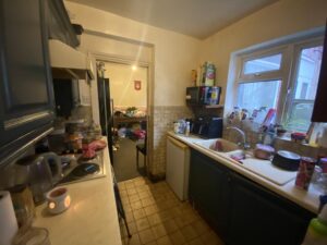 Property for sale by Auction A narrow kitchen with dark cabinets and tiled floor. Dishes are piled in a drying rack beside a sink. Small appliances like a microwave and kettle are on the counters. There's a doorway leading to a cluttered adjacent room. Presented by Palace Auctions