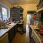 Property for sale by Auction A cluttered kitchen with dark cabinets and tiled backsplash. The countertops are filled with dishes, containers, and various kitchen items. A door in the background leads to another room, and a window lets in natural light. Presented by Palace Auctions