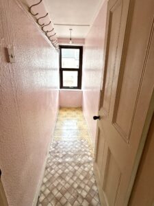 Property for sale by Auction A narrow hallway with pink textured walls and a patterned linoleum floor, leading to a window with a view of rooftops. A wooden door is partially open on the right, and several hooks are mounted along the left wall. Presented by Palace Auctions