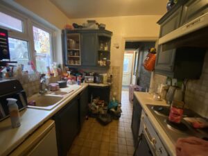 Property for sale by Auction A cluttered kitchen with dark cabinets and tiled backsplash. The countertops are filled with dishes, containers, and various kitchen items. A door in the background leads to another room, and a window lets in natural light. Presented by Palace Auctions