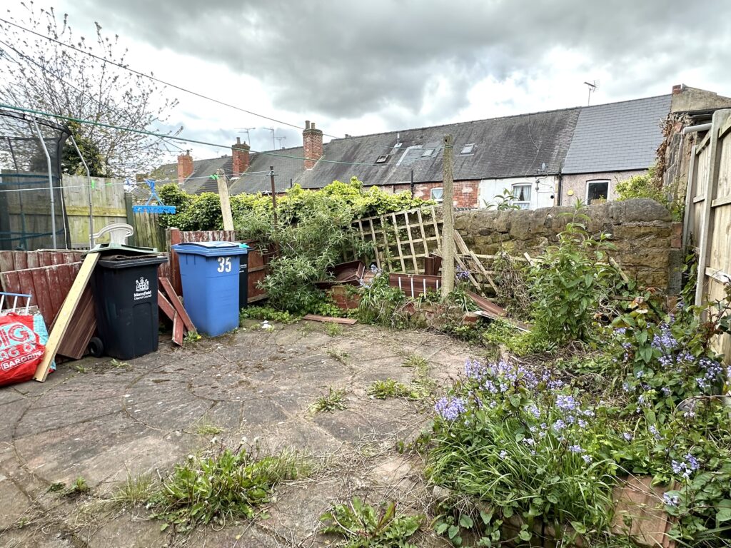 Property for sale by Auction A cluttered backyard with overgrown weeds and purple flowers. Broken wooden fences and a leaning trellis are against a stone wall. Blue and black trash bins are on the left, and brick houses with dormer windows are in the background. Cloudy sky above. Presented by Palace Auctions