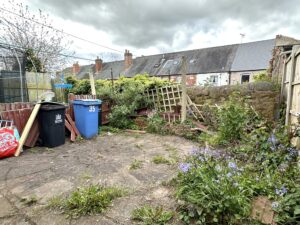 Property for sale by Auction A cluttered backyard with overgrown weeds and purple flowers. Broken wooden fences and a leaning trellis are against a stone wall. Blue and black trash bins are on the left, and brick houses with dormer windows are in the background. Cloudy sky above. Presented by Palace Auctions