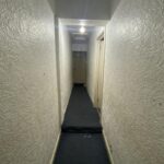 Property for sale by Auction A narrow, dimly lit hallway with textured white walls and a dark carpet. A single overhead light illuminates the space, leading to a closed door at the end of the corridor. Presented by Palace Auctions