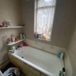 Property for sale by Auction A small bathroom with a bathtub, shelves stocked with toiletries, and a window with lace curtains. The bathtub is surrounded by various shampoo and body wash bottles. The wall paint appears worn. A bag sits on the floor nearby. Presented by Palace Auctions