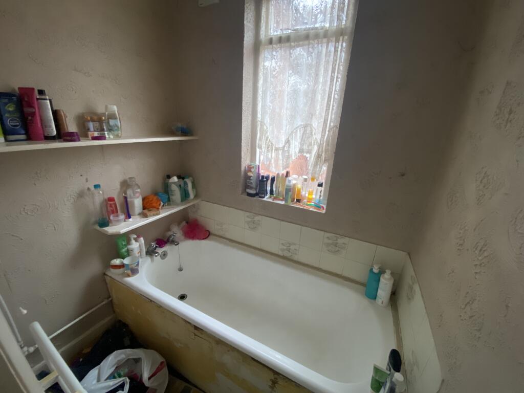 Property for sale by Auction A small bathroom with a bathtub, shelves stocked with toiletries, and a window with lace curtains. The bathtub is surrounded by various shampoo and body wash bottles. The wall paint appears worn. A bag sits on the floor nearby. Presented by Palace Auctions