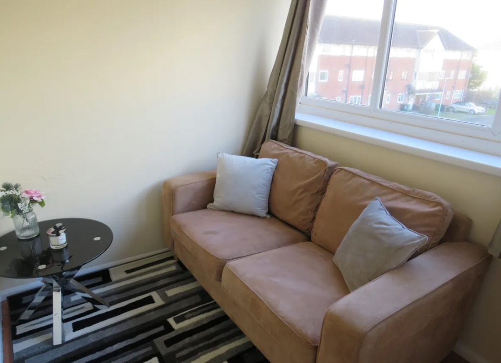 Property for sale by Auction A cozy room featuring a tan sofa with two cushions, a round black glass table with a vase of flowers and a candle, and a patterned rug. A window with brown curtains overlooks buildings outside. Presented by Palace Auctions