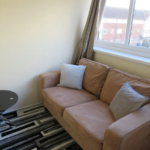 Property for sale by Auction A cozy room featuring a tan sofa with two cushions, a round black glass table with a vase of flowers and a candle, and a patterned rug. A window with brown curtains overlooks buildings outside. Presented by Palace Auctions